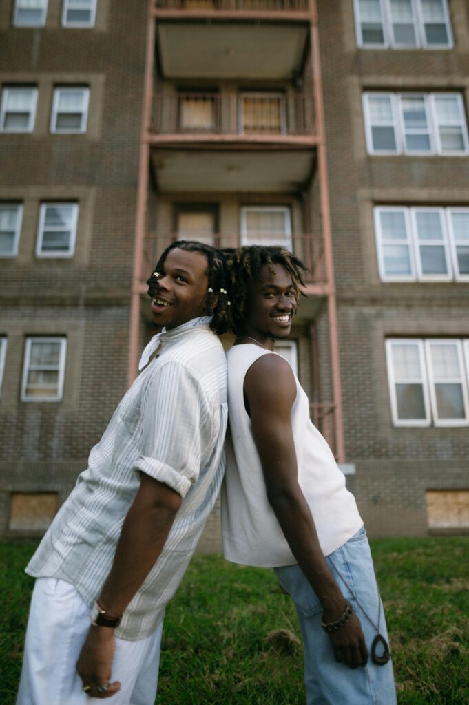 Two young men stand back to back, smiling outside an urban apartment building during summer.