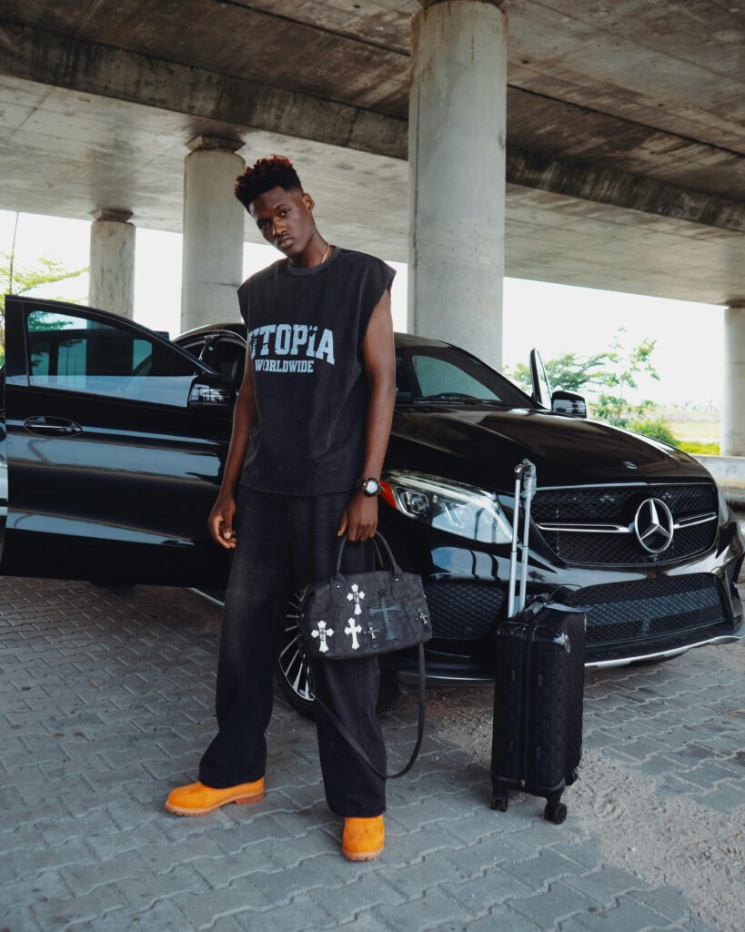 Young man in fashionable attire posing with a luxury car under a bridge.